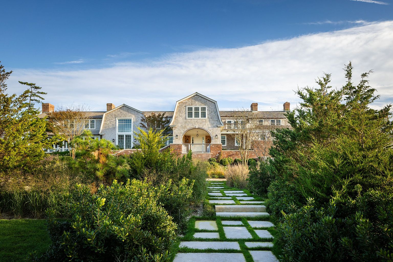 This grand shingle-style estate is captured from a low-angle perspective, emphasizing its impressive scale and symmetrical architecture. A stone-paved walkway leads through a lush, manicured garden toward a prominent arched entryway, while the house features multiple chimneys, dormer windows, and a classic cedar-shake facade. The bright blue sky and vibrant greenery create a serene and inviting atmosphere, highlighting the property's curb appeal.