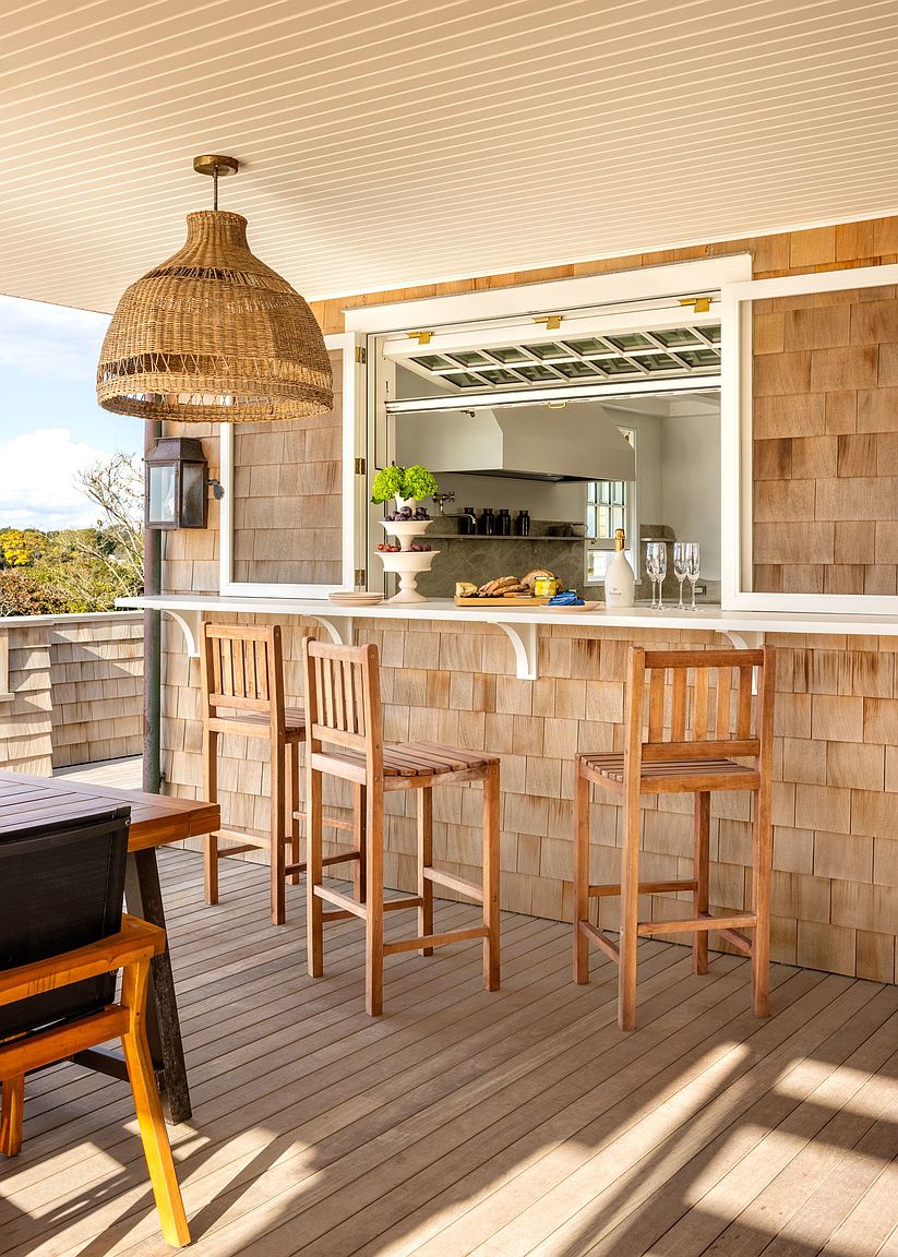 This inviting outdoor deck features a seamless transition from the kitchen through a large pass-through window, creating an ideal space for al fresco dining. The area is furnished with three wooden bar stools arranged along a built-in counter, complemented by a textured woven pendant light overhead. The exterior is finished with natural wood shingles, providing a warm, coastal aesthetic that overlooks a scenic landscape.