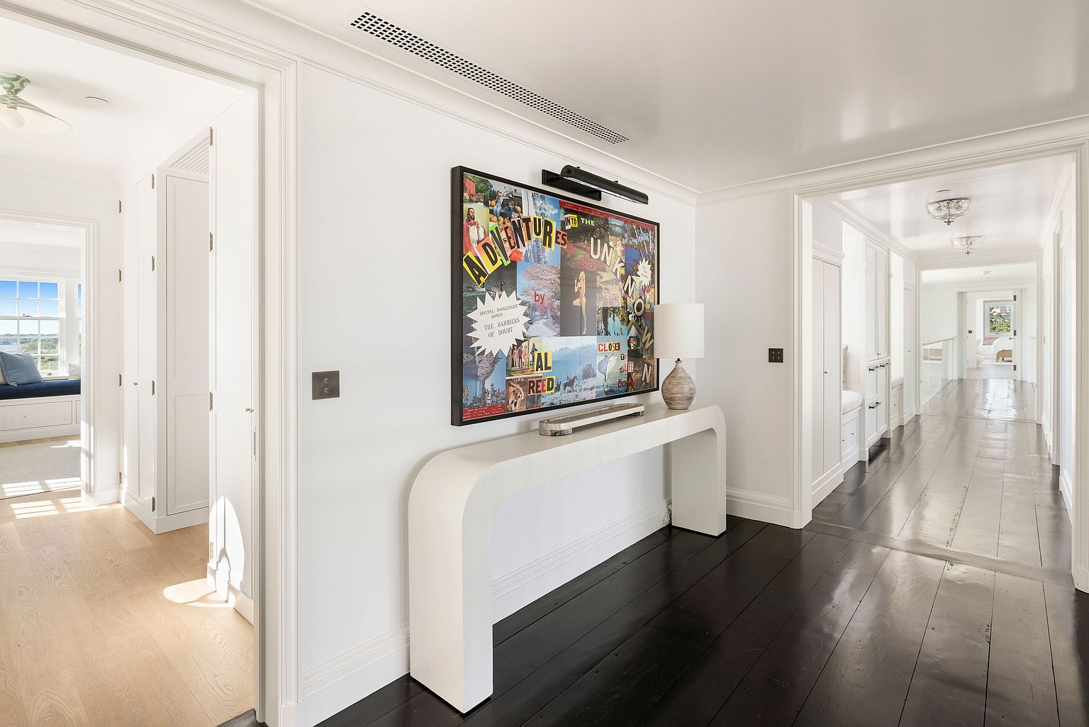 This bright and airy hallway features dark wood flooring that contrasts elegantly with crisp white walls and trim. A sleek, white console table sits beneath a large, colorful piece of framed art, which is illuminated by a dedicated picture light. The perspective looks down a long corridor, highlighting the clean architectural lines and the seamless transition between the main hallway and the adjacent rooms.