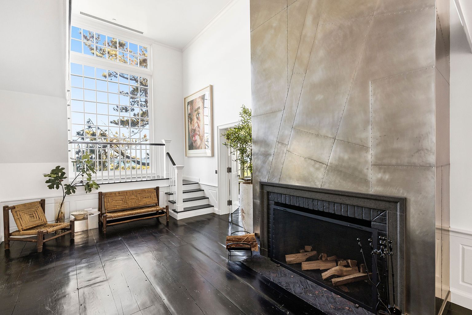 This spacious living area features dark wood flooring and a striking metal-clad chimney breast with visible rivet detailing. A large window assembly with a grid pattern allows ample natural light to fill the room, while a set of stairs leads to an elevated landing. The space is furnished with woven seating and accented by a large portrait on the wall, creating a sophisticated and artistic atmosphere.