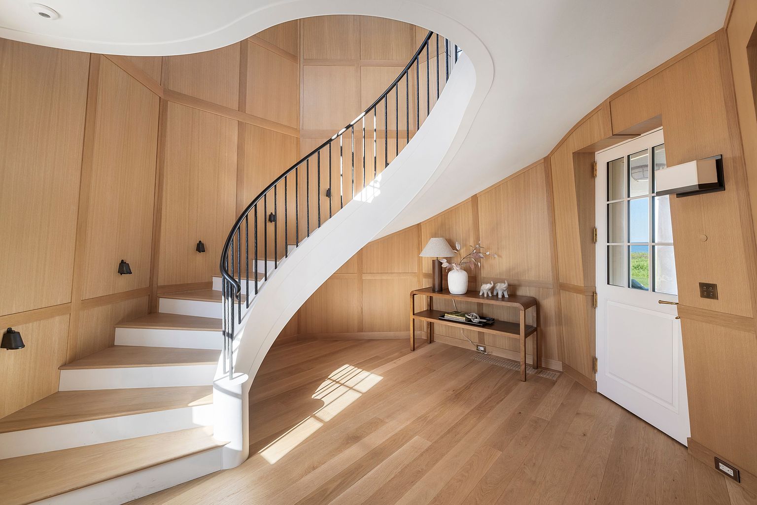 This elegant entryway features a striking curved staircase with a black metal railing and light wood treads. The walls are finished with warm-toned wood paneling, creating a cohesive and sophisticated aesthetic. A slim console table sits beneath the stairs, accented by a lamp and decorative objects, while a white door with glass panes provides a bright transition to the outdoors.