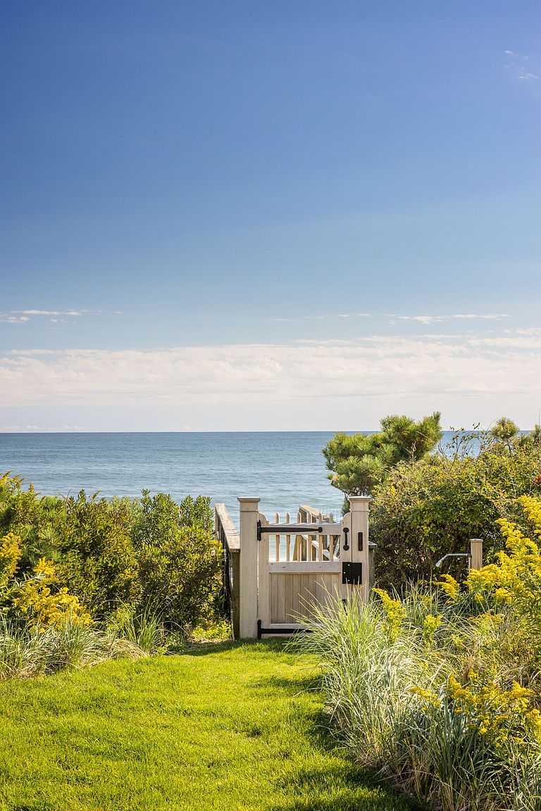 This serene outdoor space features a lush green lawn framed by vibrant yellow wildflowers and coastal greenery, leading to a white wooden gate that opens toward the ocean. The perspective is eye-level, capturing the transition from the manicured garden to the expansive blue horizon beyond the dunes. The scene evokes a peaceful, private coastal lifestyle with direct access to the beach.