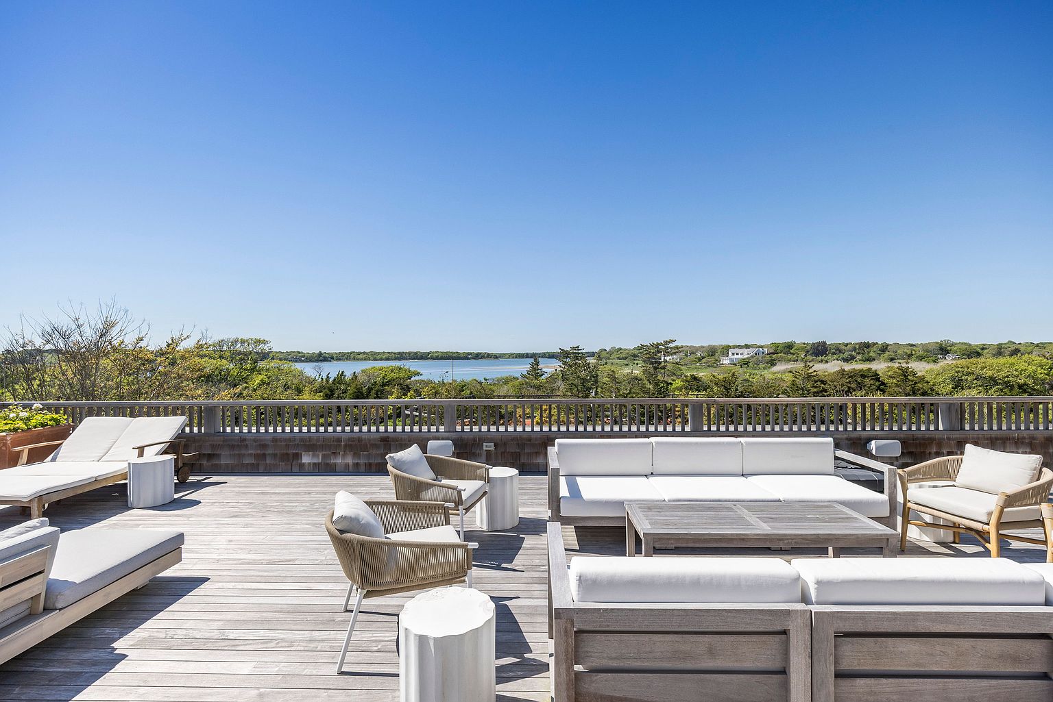 This expansive rooftop deck features a weathered wood finish and is furnished with multiple white-cushioned lounge chairs and modular seating arrangements. The space is framed by a simple wooden railing that overlooks a serene coastal landscape with a body of water in the distance. The bright, clear sky and open layout create an inviting atmosphere perfect for outdoor relaxation and entertaining.