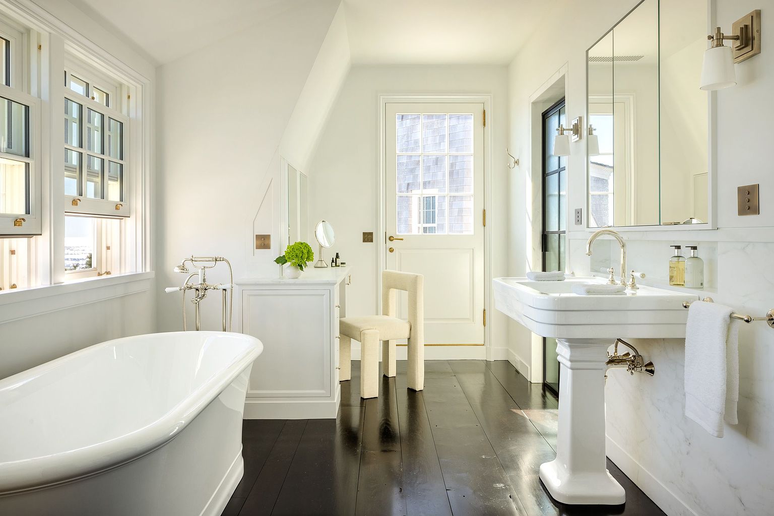 This bright and airy primary bathroom features a classic freestanding soaking tub positioned near large windows, complemented by a pedestal sink and a dedicated vanity area with a modern chair. The space is defined by its clean white walls, dark wood flooring, and a door leading to an exterior space, creating a sophisticated and serene atmosphere. The perspective is a wide-angle shot that captures the entire layout, emphasizing the elegant fixtures and the seamless blend of traditional and contemporary design elements.