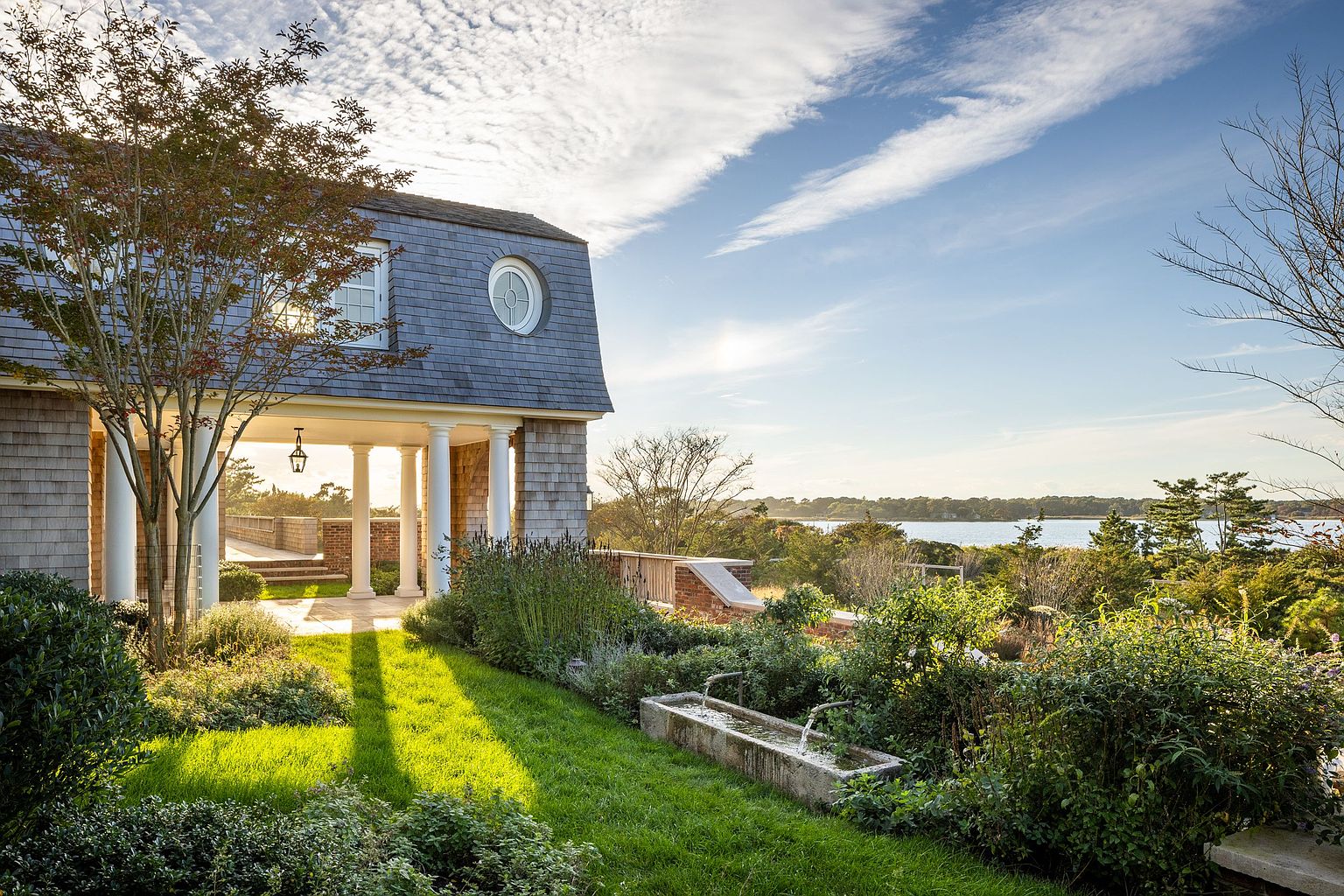 This serene outdoor scene captures a shingled home with a classic gambrel roof and a covered porch supported by white columns. In the foreground, a stone water feature sits amidst lush greenery and a manicured lawn, leading the eye toward a scenic waterfront vista. The golden hour lighting enhances the peaceful, coastal atmosphere of this well-landscaped property.