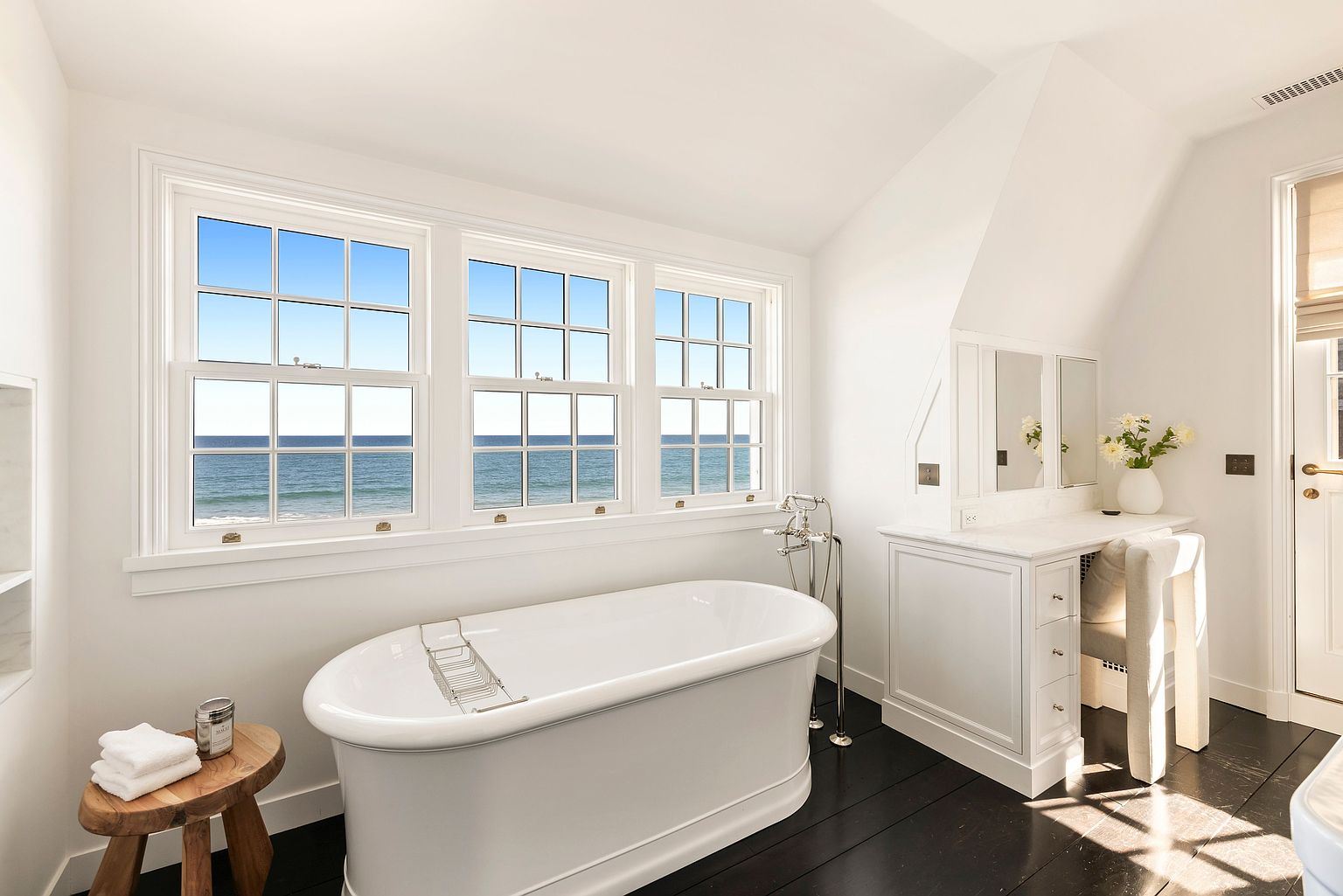This bright and airy primary bathroom features a classic freestanding soaking tub positioned beneath a trio of large windows that offer expansive ocean views. To the right, a built-in vanity with a stone countertop and integrated mirrors provides a dedicated space for grooming, complemented by a light-toned chair. The room is finished with dark wood flooring, creating a sophisticated contrast against the crisp white walls and trim.