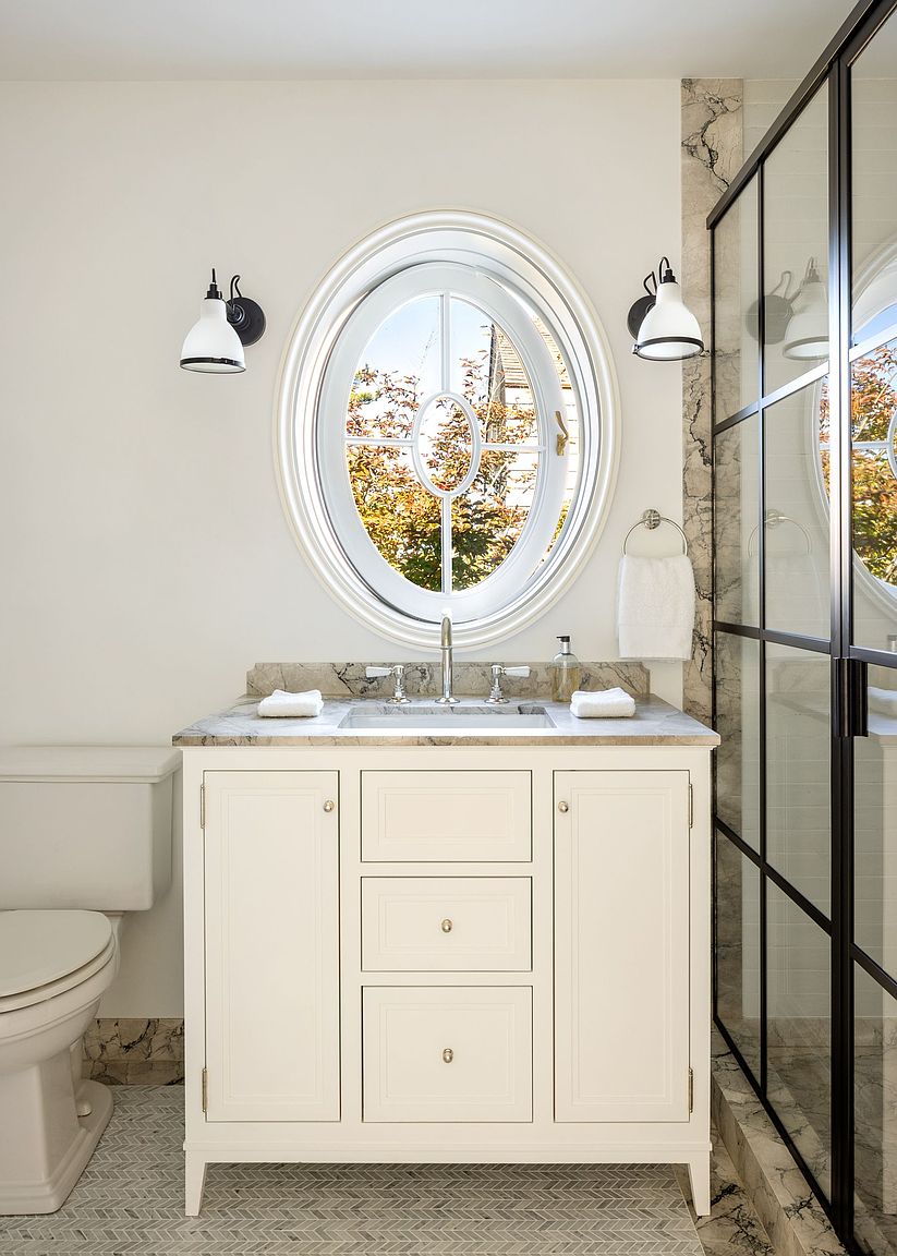 This bright bathroom features a central vanity with a stone countertop and a white cabinet base, positioned beneath a distinctive oval window. To the right, a glass-enclosed shower with a dark metal frame adds a modern, industrial touch, while the floor is finished in a subtle herringbone-patterned tile. The space is illuminated by two wall-mounted sconces, creating a clean and sophisticated aesthetic.