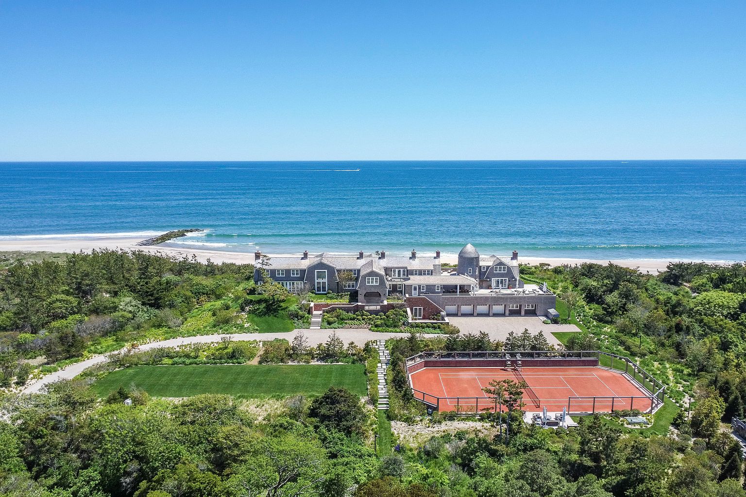 This expansive aerial view captures a luxurious coastal estate nestled along a pristine shoreline. The property features a sprawling shingle-style residence, a private red clay tennis court, and a manicured lawn, all surrounded by lush coastal vegetation. The cinematic perspective highlights the seamless integration of the home with its natural beachfront environment under a clear, bright sky.