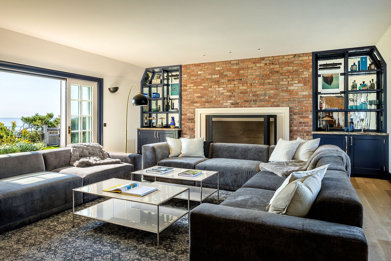 This spacious living room features a large, plush gray sectional sofa arranged around two matching glass-topped coffee tables on a patterned area rug. A prominent brick wall serves as a focal point, housing a fireplace with a simple surround, flanked by dark-painted built-in shelving units. Large glass doors to the left provide abundant natural light and a scenic view of the outdoor landscape, creating an inviting and comfortable atmosphere.