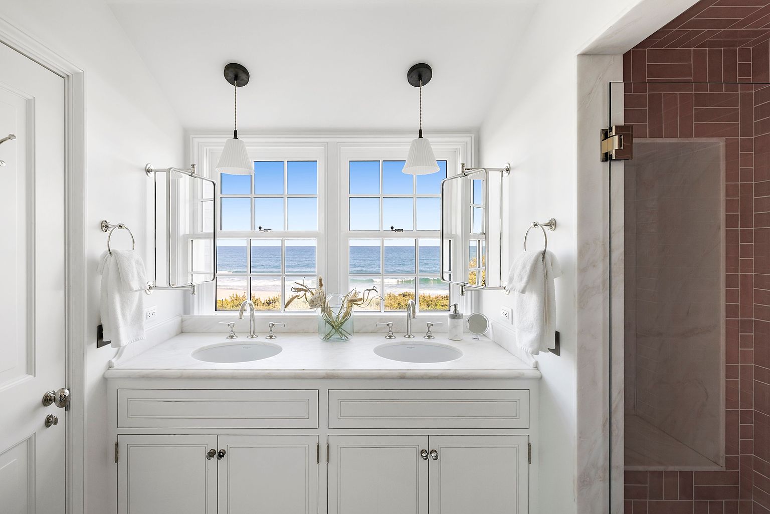 This bright and airy primary bathroom features a double vanity with a stone countertop and white cabinetry, positioned directly in front of a large window with a stunning ocean view. The space is accented by two pendant lights, wall-mounted mirrors, and white towels hanging on either side of the vanity. To the right, a glimpse of a shower area with reddish-brown rectangular wall tiles adds a warm contrast to the crisp, white aesthetic of the room.