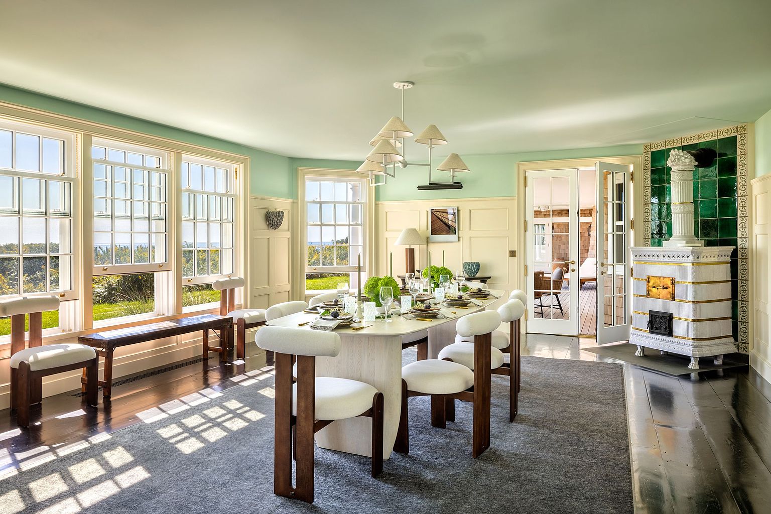 This elegant dining room features a long, light-toned table surrounded by modern chairs with dark wood frames and white upholstery. The space is bathed in natural light from large windows that offer views of the outdoors, while a unique, tall, decorative stove with green tile accents serves as a striking focal point. The room is finished with light green walls, wainscoting, and a dark wood floor covered by a large, neutral-toned area rug.