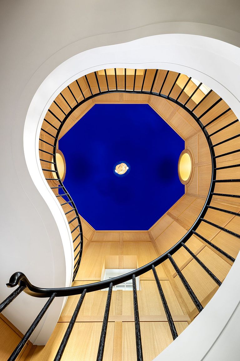 This dramatic low-angle shot captures a spiral staircase looking upward toward a striking, vibrant blue octagonal ceiling feature. The staircase features a sleek black metal railing that curves elegantly against light wood-paneled walls, creating a sophisticated and modern architectural aesthetic. The perspective emphasizes the verticality and geometric interplay between the curved stairs and the angular ceiling design.