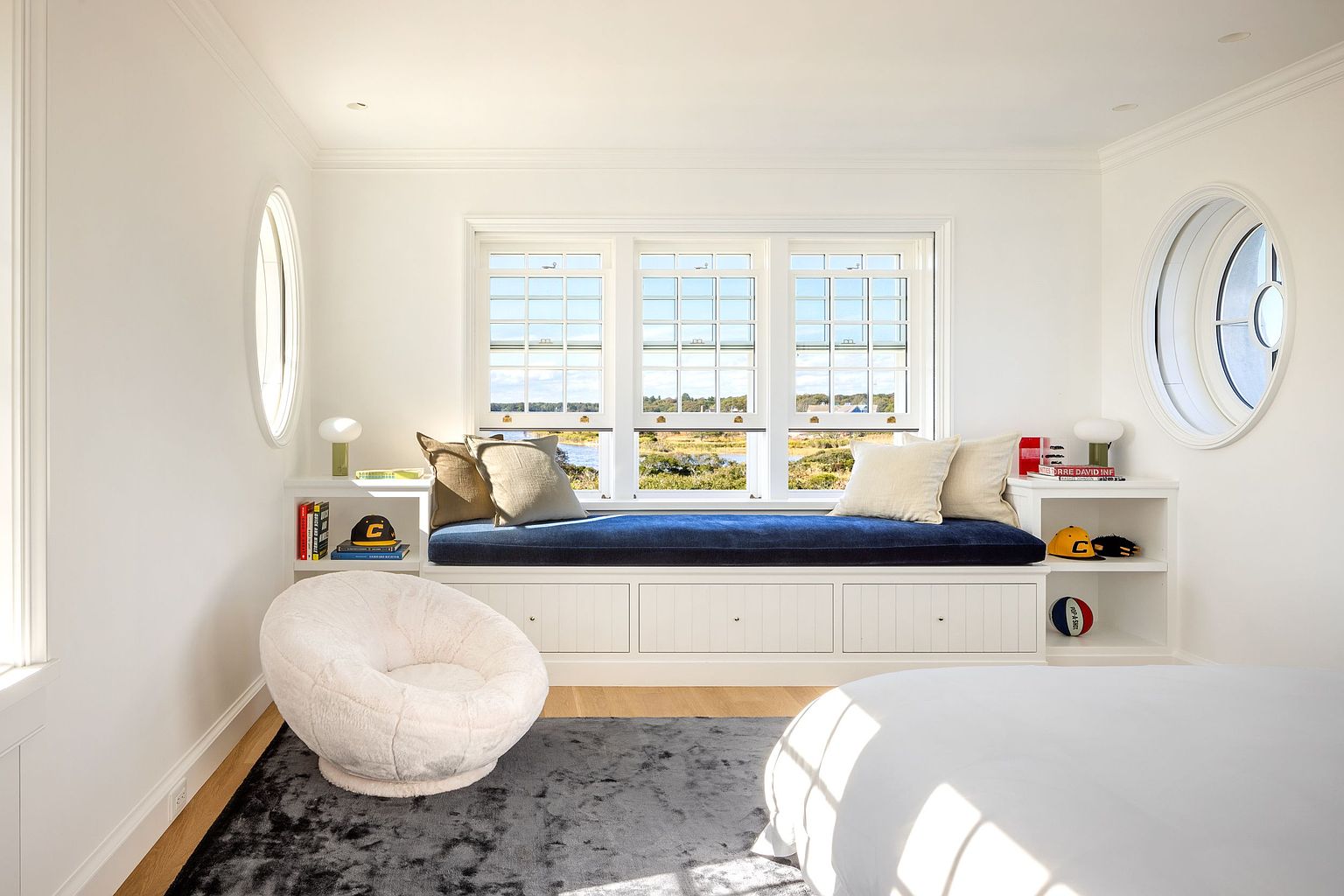 This bright and airy bedroom features a prominent built-in window seat with deep blue upholstery, flanked by open shelving and unique oval-shaped accent windows. A plush, cream-colored circular chair sits on a textured gray rug, creating a cozy reading nook beneath the large central window arrangement. The room is finished with crisp white walls and clean trim, offering a serene and well-lit atmosphere.