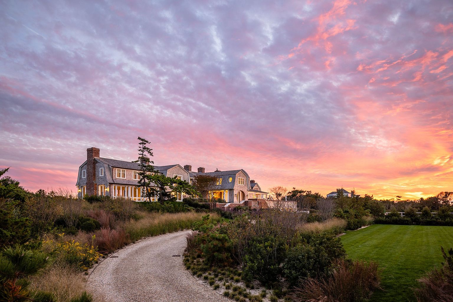 This stunning shingle-style estate is captured at sunset, showcasing a sprawling residence nestled within a lush, natural landscape. A winding gravel driveway leads toward the home, which features multiple chimneys, dormer windows, and a warm, inviting glow emanating from the interior lights. The dramatic, vibrant pink and orange sky provides a cinematic backdrop to the manicured lawn and native grasses in the foreground.
