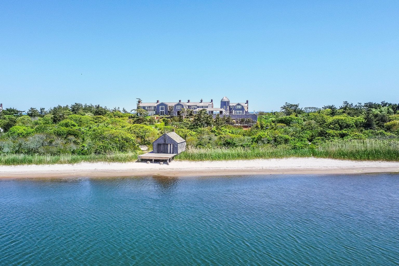 This aerial perspective captures a sprawling, shingle-style estate nestled atop a lush, tree-covered coastal bluff. In the foreground, a weathered wooden boathouse sits directly on the sandy shoreline, connected to a small deck that extends toward the calm, deep blue water. The scene conveys a sense of secluded luxury and serene waterfront living, framed by a clear, expansive sky.