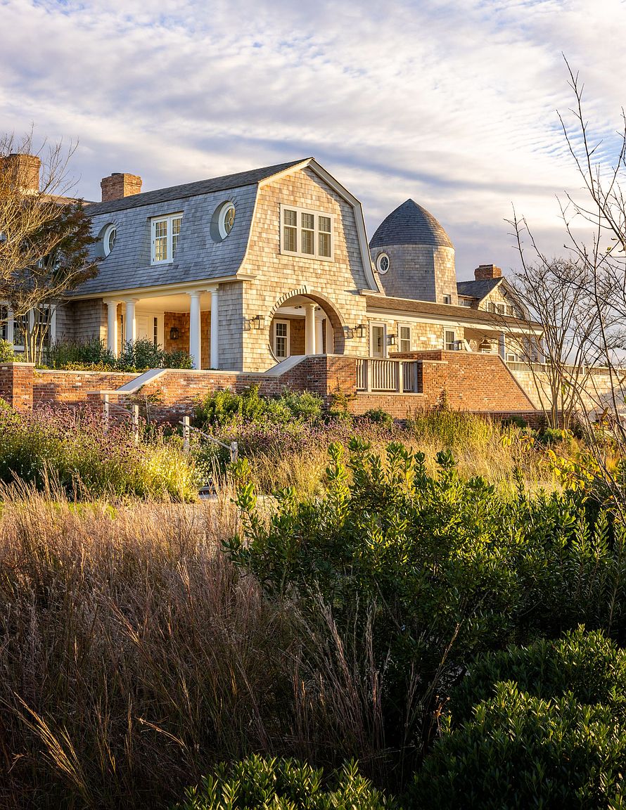 This elegant shingle-style estate features a prominent gambrel roofline, a distinctive circular tower, and a grand arched entryway supported by classic columns. The home is elevated on a substantial brick foundation, overlooking a lush, naturalistic landscape filled with tall grasses and native plantings. The warm, golden-hour light enhances the textured wood siding and brickwork, creating a sophisticated and inviting coastal atmosphere.