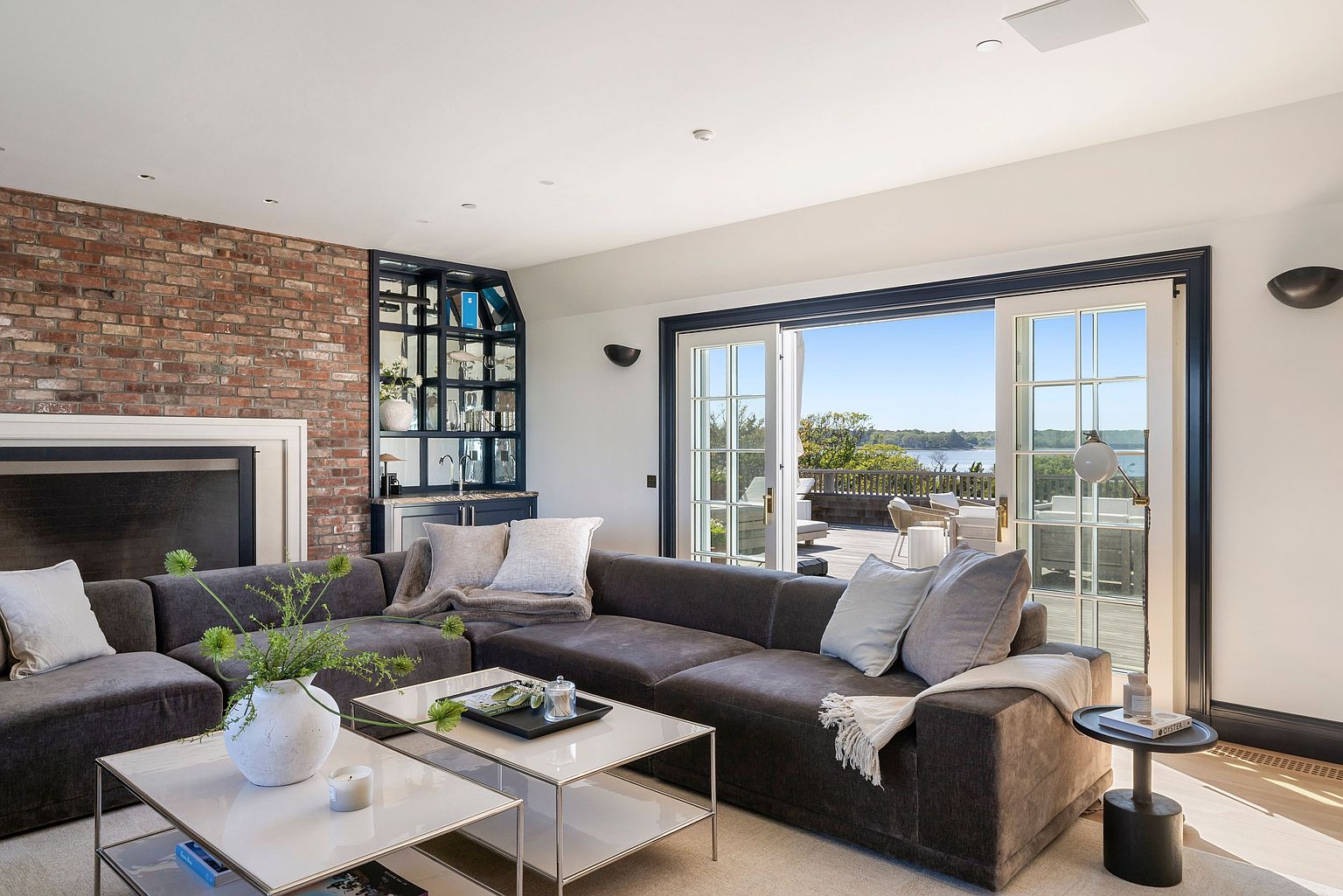 This sophisticated living room features a large, dark-toned sectional sofa arranged around a pair of modern, glass-topped coffee tables. A prominent brick chimney breast anchors the space, complemented by a built-in dark-toned bar area with glass shelving. Large French doors open onto a deck, providing a seamless transition to the outdoors and a scenic water view.