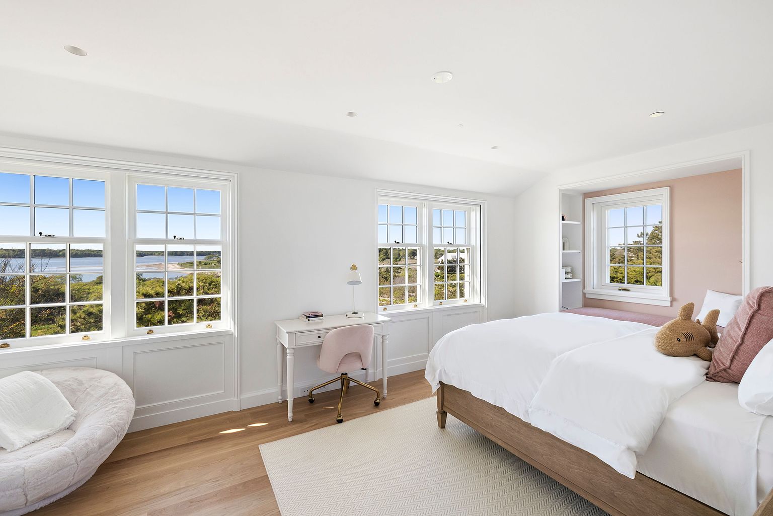 This bright and airy bedroom features light wood flooring, white walls, and a large bed with crisp white linens. A dedicated desk area with a pink chair sits between two sets of windows that offer expansive water views, while a cozy nook with a built-in bench and soft pink accent wall provides a comfortable reading spot. The room is decorated with a plush circular chair and a stuffed animal, creating a serene and inviting atmosphere.