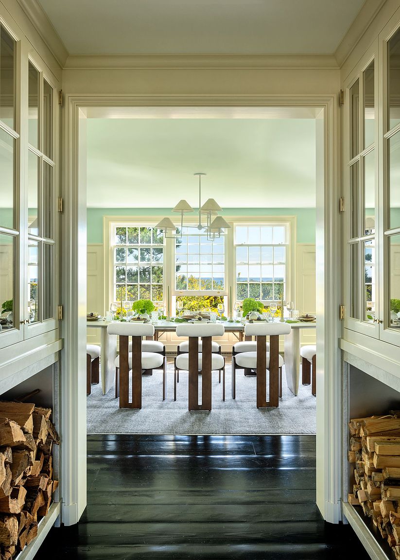 This view captures a formal dining room framed by a transitionary hallway featuring built-in cabinetry with integrated firewood storage at the base. The dining area is bathed in natural light from a trio of large windows, showcasing a long table with modern chairs and a minimalist chandelier. The contrast between the dark wood flooring in the foreground and the light, airy aesthetic of the dining space creates a sophisticated and inviting atmosphere.