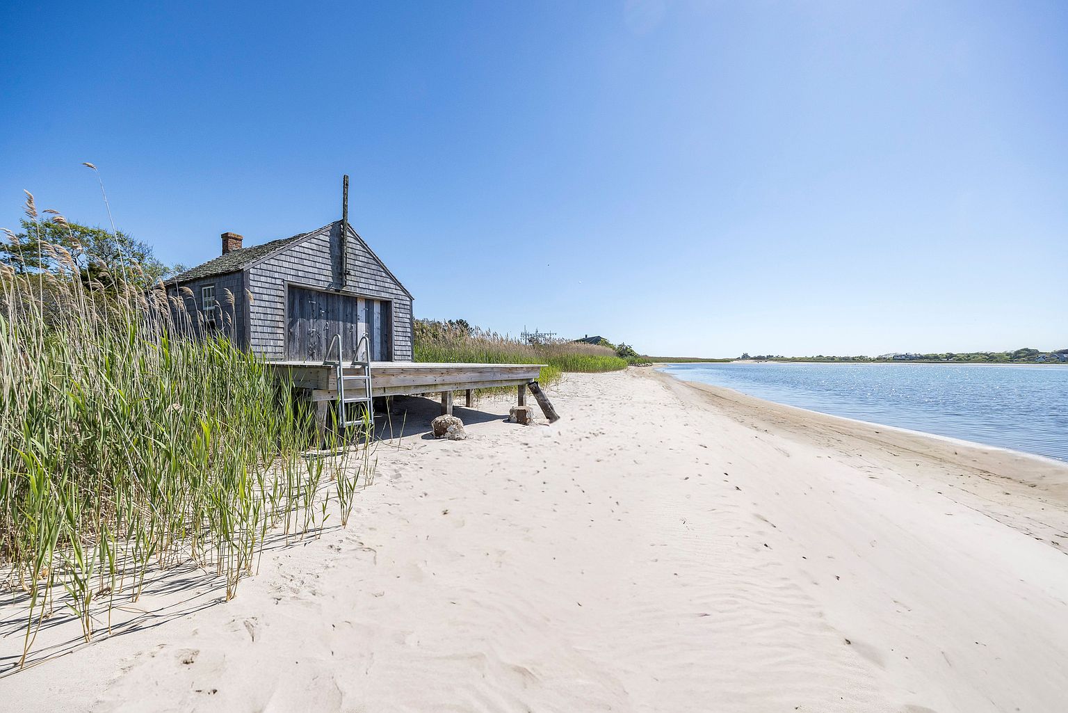 This image captures a weathered, shingled coastal structure situated directly on a sandy beach. A wooden deck extends from the building toward the water, featuring a metal ladder for easy access to the shoreline. The scene is framed by tall beach grasses in the foreground and a clear, expansive blue sky, evoking a serene and secluded waterfront atmosphere.