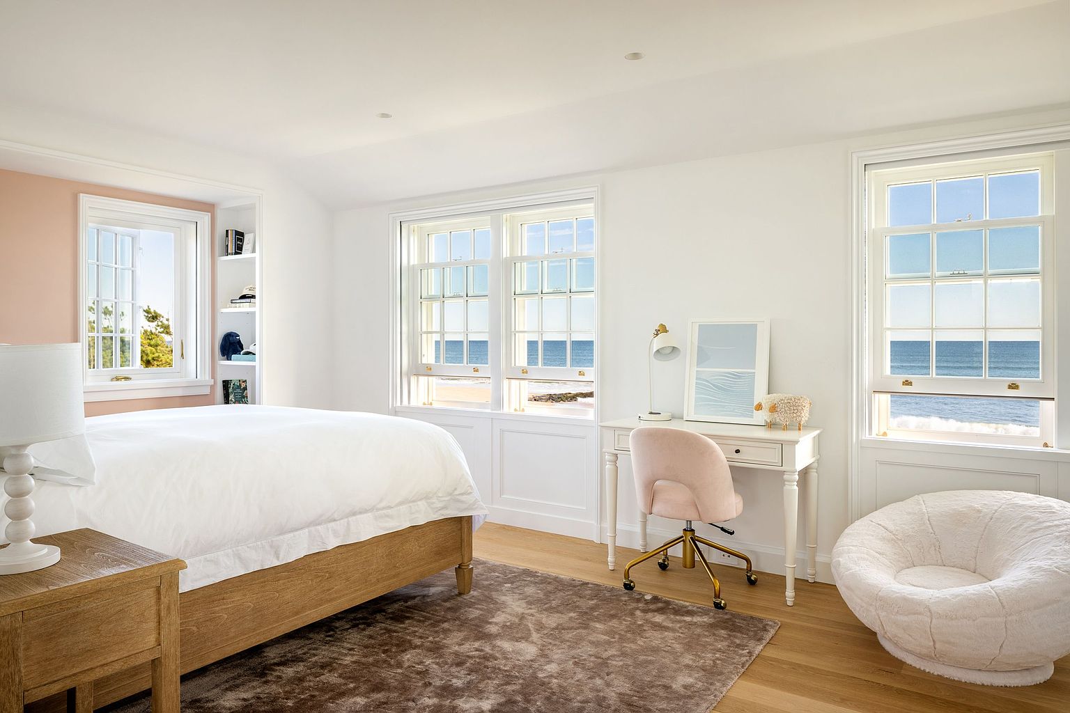 This bright and airy bedroom features a light wood bed frame with crisp white bedding, complemented by a soft pink accent wall and a plush, textured area rug. A dedicated workspace with a desk and a soft pink chair is positioned near large windows that offer expansive, unobstructed views of the ocean. The room is finished with a cozy, round white chair and built-in shelving, creating a serene and inviting coastal retreat.