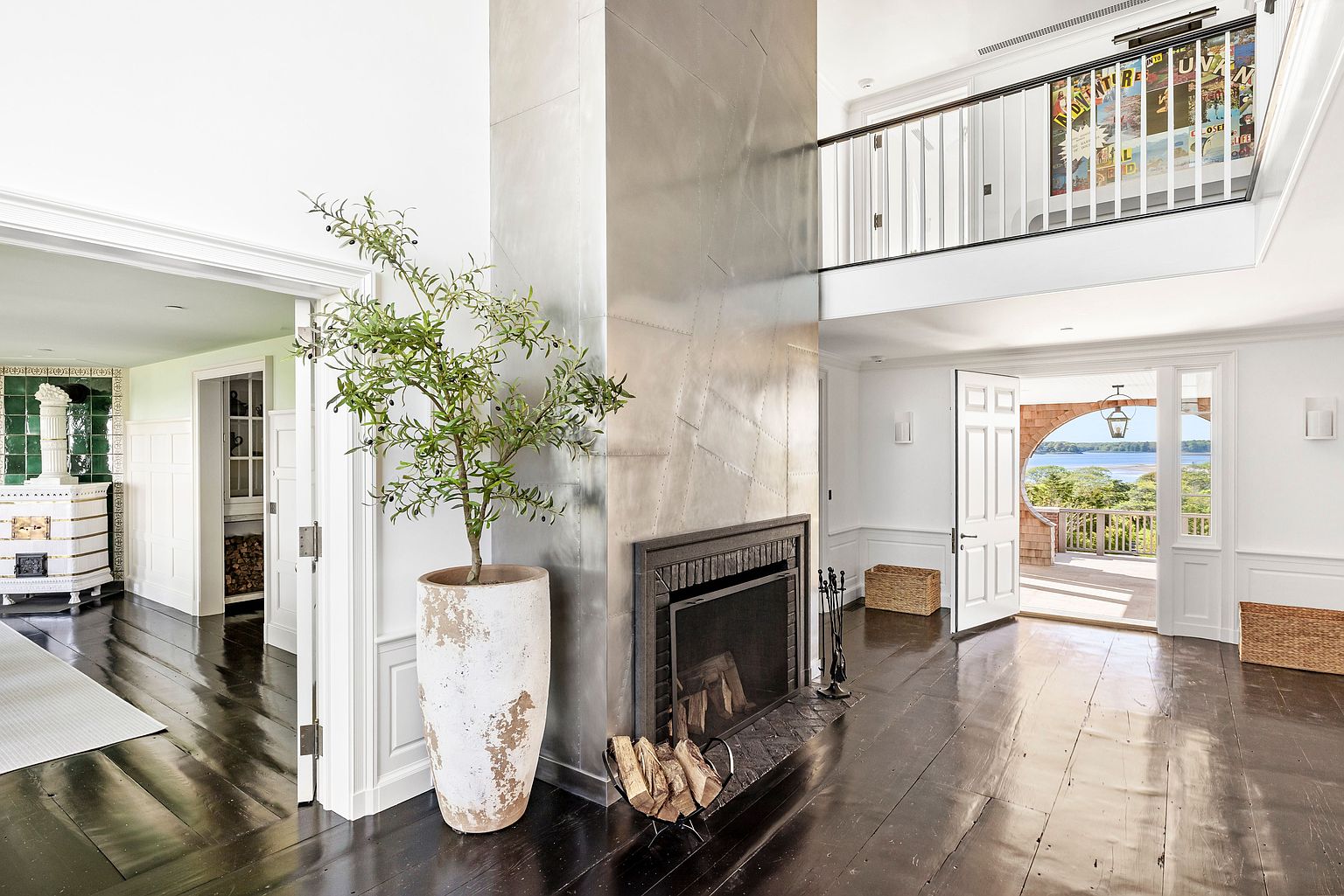 This spacious living area features a striking metal-clad chimney breast that serves as a central focal point, complemented by dark wood flooring throughout. A large potted olive tree adds a touch of greenery, while an open doorway leads to a bright, airy space with a circular window overlooking a scenic waterfront view. The room connects to an upper-level mezzanine, creating an open and expansive atmosphere with a blend of industrial and traditional design elements.