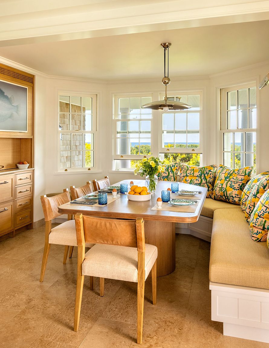 This bright and inviting dining area features a custom built-in corner banquette adorned with vibrant, patterned cushions, paired with a light wood dining table and matching chairs. Large windows wrap around the space, offering a scenic view of the outdoors and flooding the room with natural light. A stylish pendant light hangs above the table, creating a warm and cohesive atmosphere perfect for casual family meals.