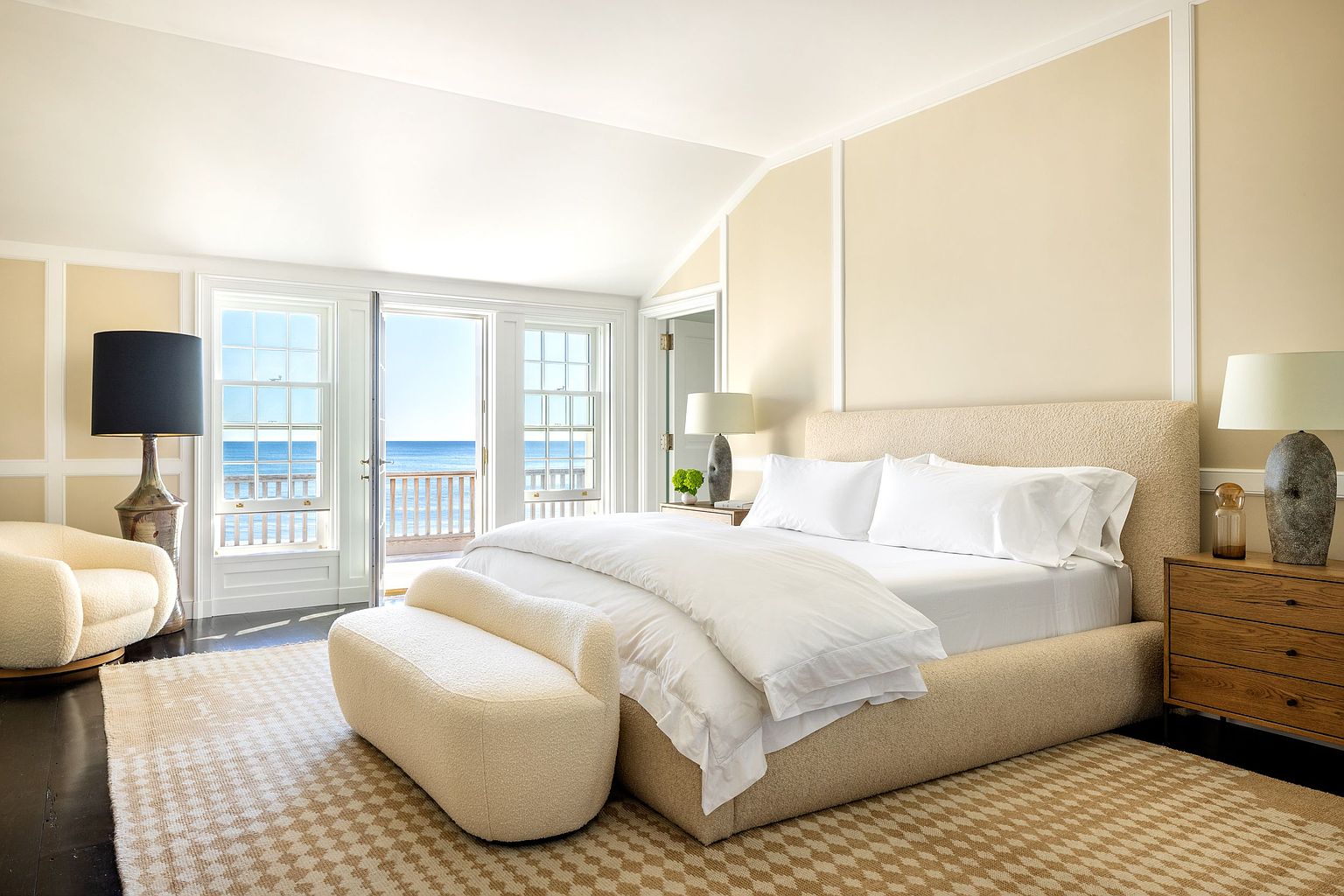 This serene primary bedroom features a plush, textured bed frame and matching bench, complemented by a neutral color palette and a patterned area rug. Large glass doors open directly to a private deck with expansive ocean views, flooding the room with natural light. The space is accented by warm wood nightstands and sculptural table lamps, creating a sophisticated and tranquil coastal retreat.