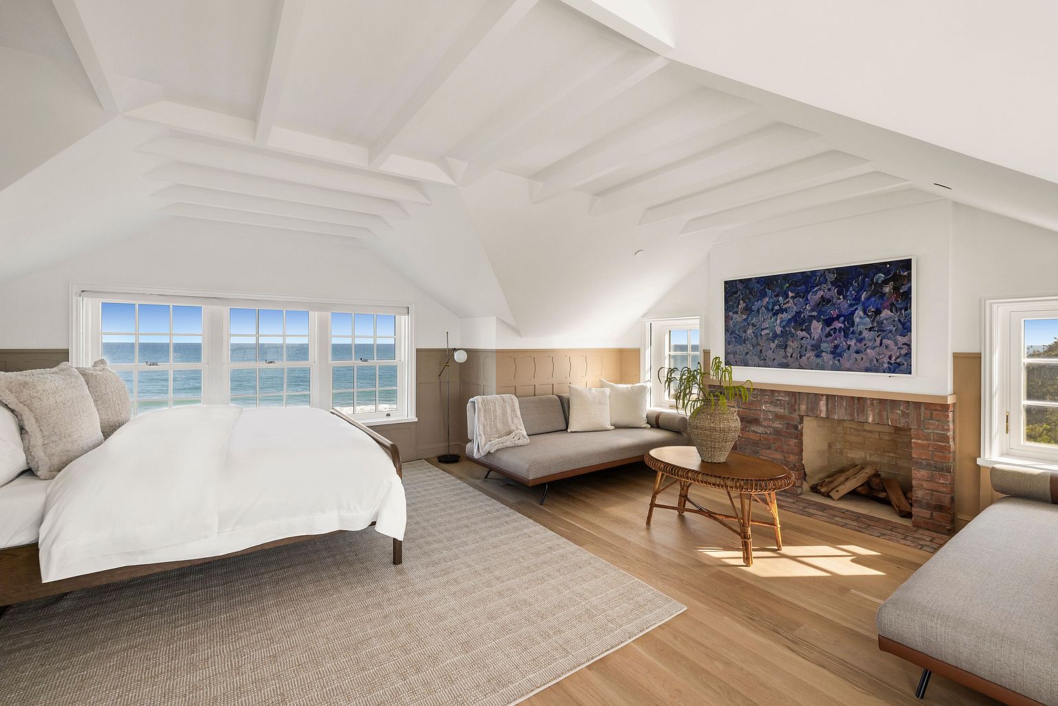 This bright and airy primary bedroom features vaulted ceilings with exposed beams and a large window bank offering expansive ocean views. The room is furnished with a comfortable bed, two modern daybeds, and a central coffee table, all arranged around a brick fireplace with a prominent abstract painting above it. The space is finished with light wood flooring and a neutral color palette, creating a serene and sophisticated coastal retreat.