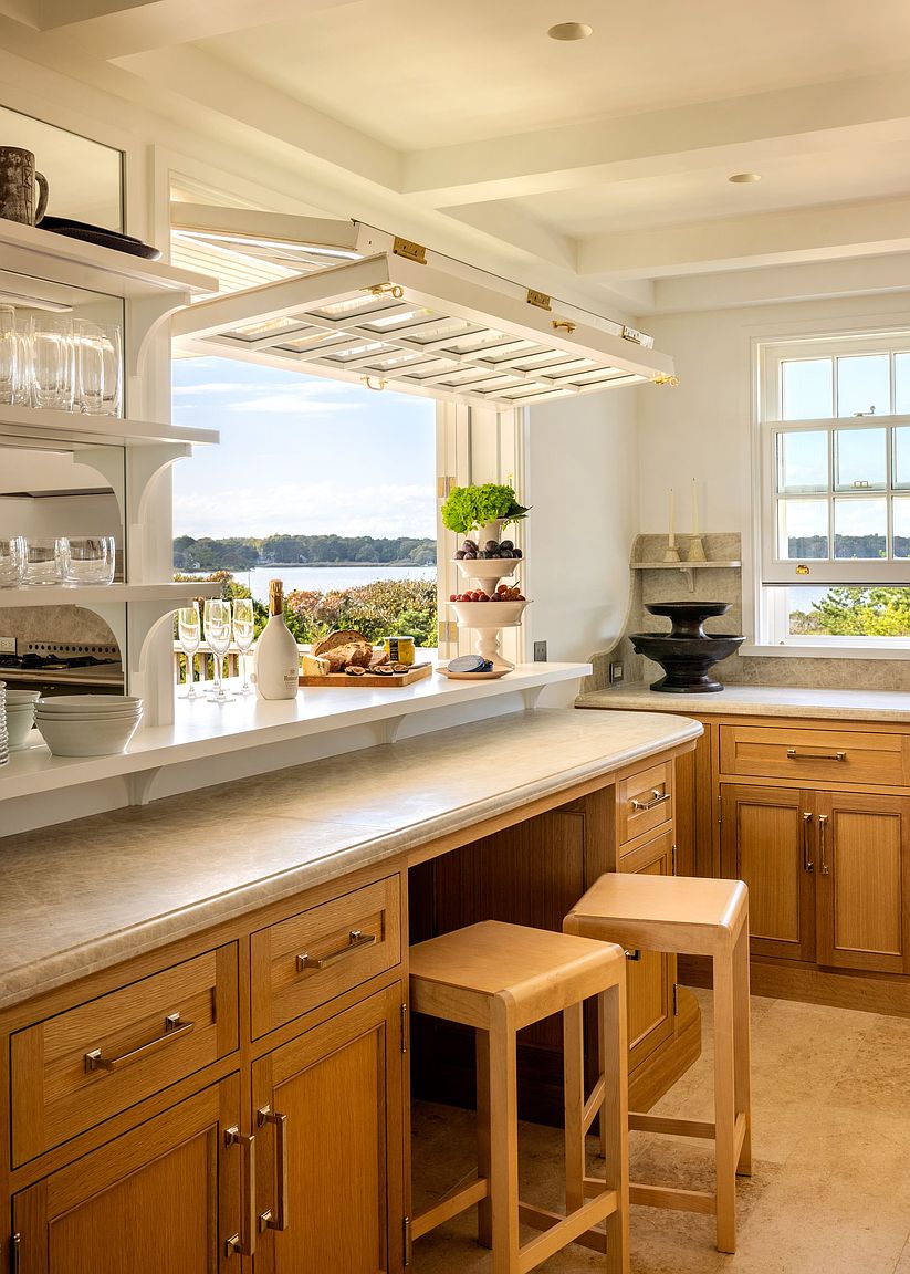 This bright kitchen features a pass-through window that opens upward to reveal a scenic waterfront view, seamlessly connecting the indoor space with the outdoors. The area includes a light wood breakfast bar with matching stools, open shelving for glassware, and stone countertops. The overall aesthetic is airy and coastal, emphasizing natural light and a functional, open-concept layout.