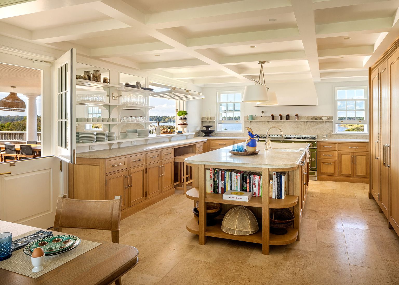 This bright and airy kitchen features light wood cabinetry, a large central island with integrated bookshelves, and stone countertops. The space is defined by a coffered ceiling and a pass-through window that opens to an outdoor deck, creating a seamless indoor-outdoor flow. A professional-style range and neutral stone flooring complete the sophisticated, coastal-inspired design.