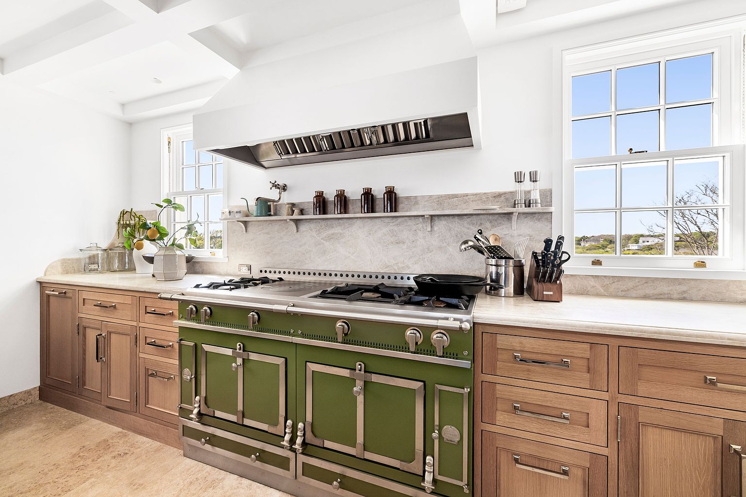 This high-end kitchen features a prominent, professional-grade green range with a matching stainless steel hood, serving as the focal point of the space. The room is equipped with light wood cabinetry, stone countertops, and a stone backsplash, complemented by two windows that provide natural light and views of the outdoors. The perspective is a straight-on eye-level shot, highlighting the clean, classic design and premium culinary appliances.