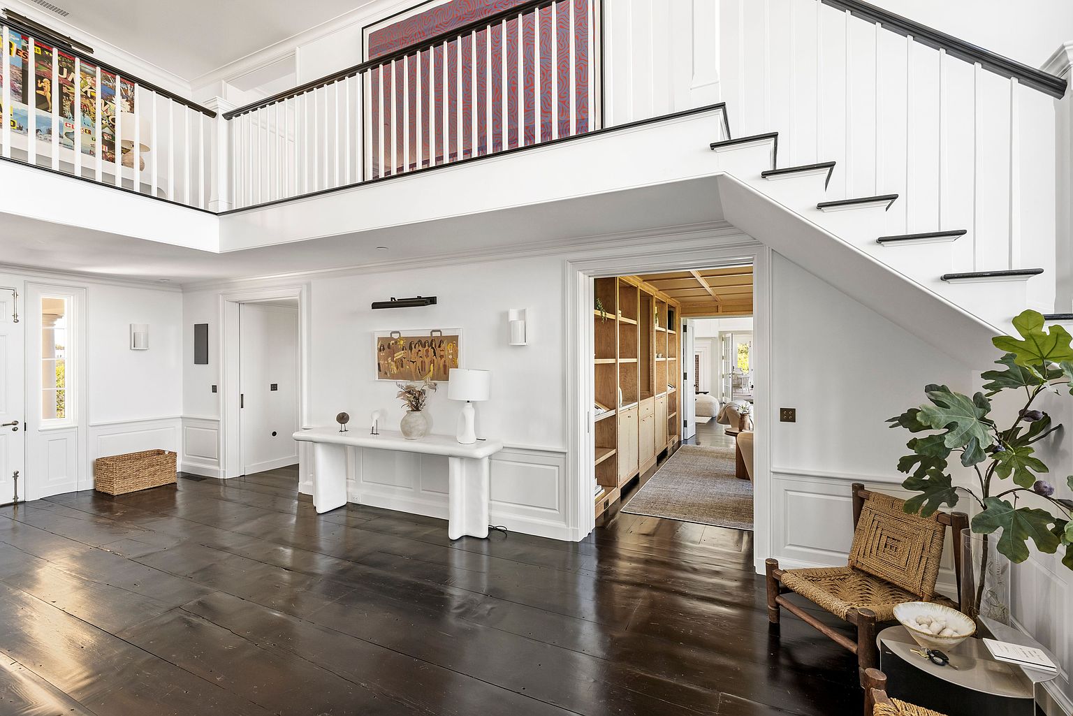 This elegant entryway features a dramatic staircase with a black handrail and white balusters, leading to an upper landing. The space is anchored by dark, wide-plank wood flooring and a minimalist white console table adorned with a lamp and decorative accents. A doorway leads into a warm, wood-paneled library or study, while a large piece of abstract art hangs above the landing, creating a sophisticated and airy atmosphere.