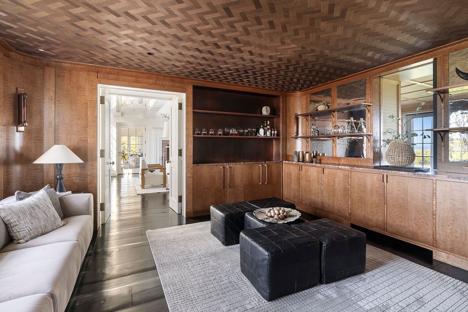 This sophisticated study or lounge area features rich wood-paneled walls and a distinctive herringbone-patterned wood ceiling. The space is furnished with a neutral sofa, four black leather ottomans arranged as a central coffee table, and extensive built-in cabinetry with open shelving. A doorway provides a view into a bright, airy adjacent room, creating a sense of depth and architectural continuity.