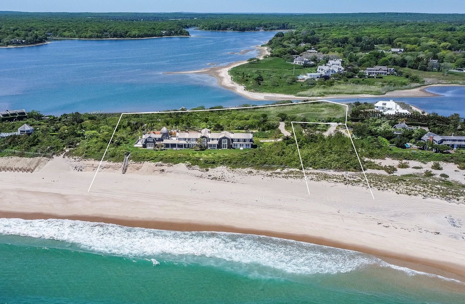 This high-angle aerial view captures a sprawling, shingle-style estate situated on a private coastal peninsula. The property features a long, multi-sectioned residence nestled between a pristine sandy beach and a serene, winding inlet. The expansive grounds include natural coastal landscaping, a private staircase leading to the shore, and a clearly defined property boundary outlined in white.