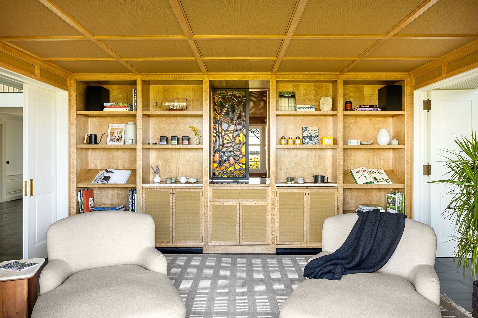 This sophisticated living space features a symmetrical arrangement of two plush, light-toned lounge chairs facing a large, custom-built light wood shelving unit. The shelving unit incorporates a central stained-glass panel, woven cabinet door inserts, and open display shelves filled with books and decorative objects. A coffered ceiling with a textured finish and a geometric-patterned area rug complete the refined, tranquil aesthetic of the room.