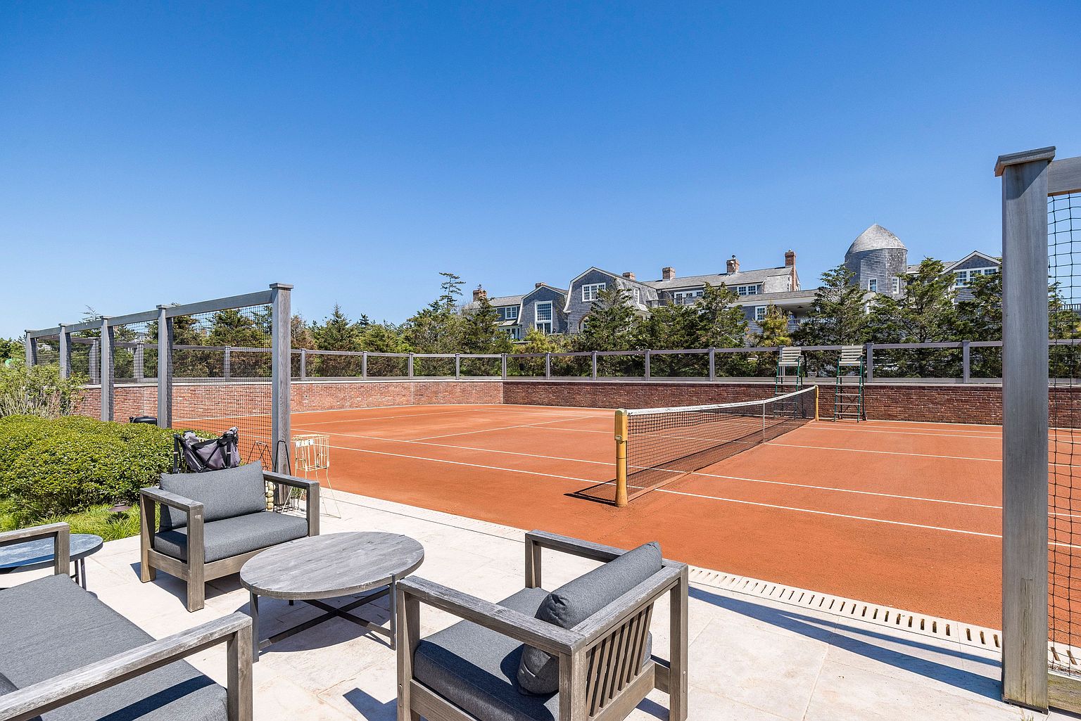 This image captures a well-maintained clay tennis court situated adjacent to a comfortable outdoor lounge area. The foreground features modern wooden patio furniture with plush gray cushions, while the court is enclosed by a brick wall and protective netting. In the background, a large, multi-story shingled building with a distinctive turret adds a classic coastal architectural aesthetic to the scene.
