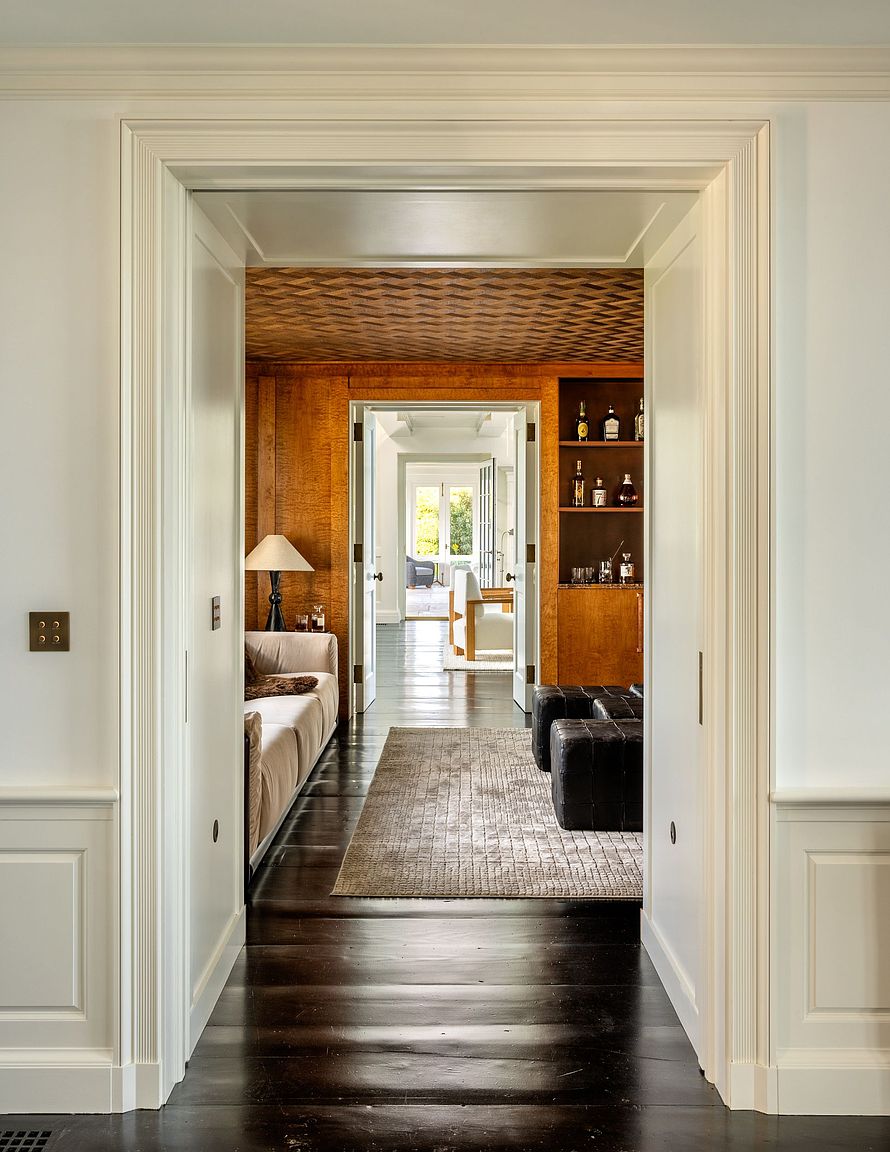This perspective captures a series of enfilade rooms, looking through a white-trimmed doorway into a warm, wood-paneled study and beyond to a bright sunlit room. The foreground features dark wood flooring and white wainscoting, while the middle room showcases a unique geometric patterned ceiling and a built-in bar area. The composition emphasizes depth and architectural layering, creating a sophisticated and inviting transition between living spaces.