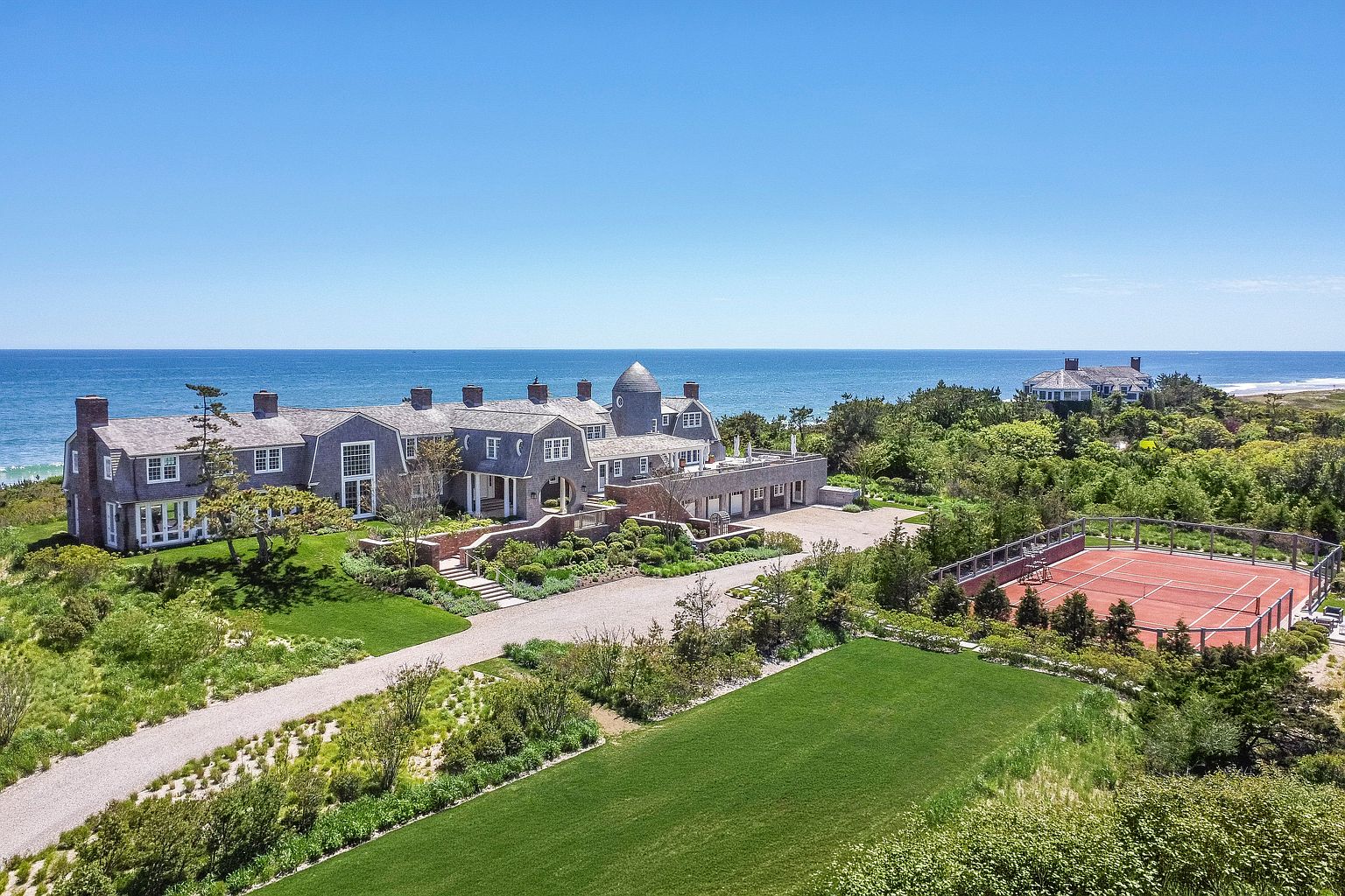 This high-angle aerial view captures a sprawling, shingle-style coastal estate situated on a lush, grassy bluff overlooking the ocean. The property features a large main residence with a distinctive turret, a private clay tennis court, and extensive landscaped grounds with gravel pathways. The cinematic perspective emphasizes the expansive waterfront location and the seamless integration of the architecture with the surrounding natural coastal environment.