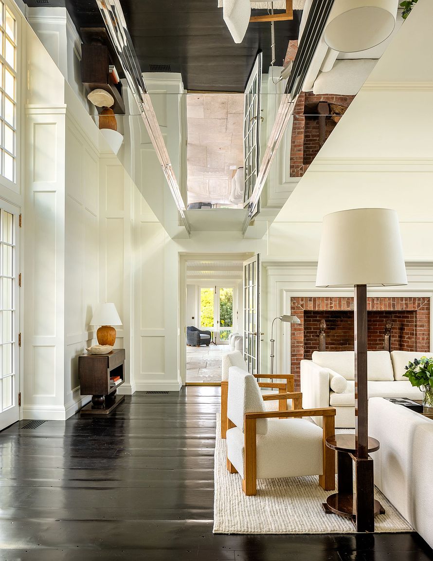 This elegant living room features dark wood flooring and a striking mirrored ceiling panel that reflects the space below. A brick fireplace serves as a focal point, complemented by light-toned upholstered seating and a tall floor lamp. The room flows into an adjacent sunlit hallway, creating an airy and sophisticated atmosphere.