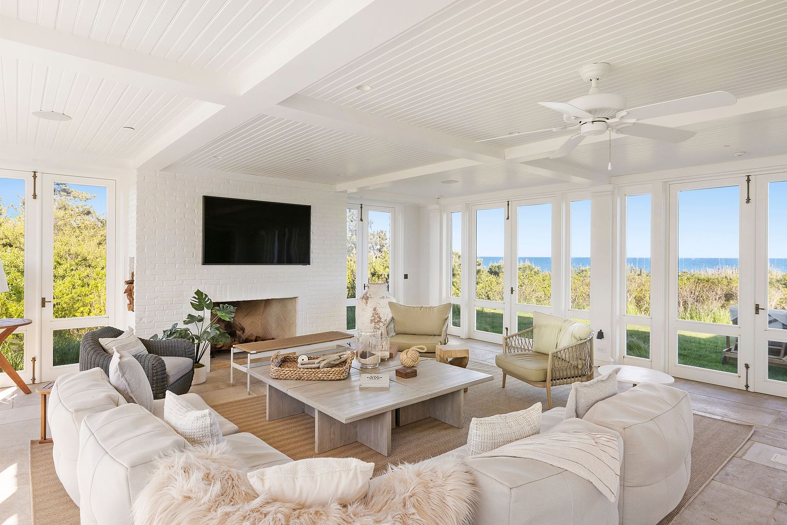 This bright and airy living room features a large, plush white sectional sofa arranged around a square light wood coffee table. A white brick wall houses a fireplace, while expansive windows and glass doors provide panoramic views of the ocean and surrounding greenery. The space is finished with a white paneled ceiling, a ceiling fan, and a neutral area rug, creating a relaxed, coastal-inspired atmosphere.