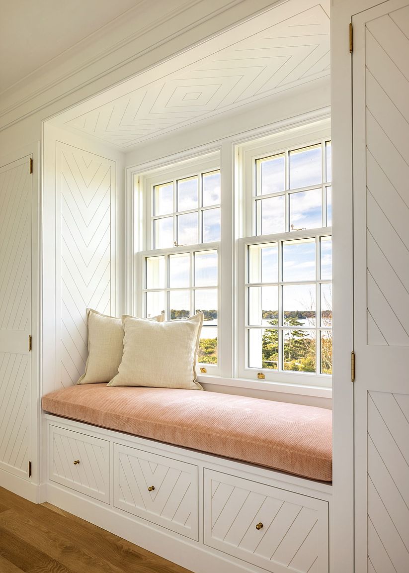 This image showcases a custom-built window seat nook framed by white cabinetry with a sophisticated herringbone pattern. The bench features a soft, textured cushion in a muted peach tone, complemented by two neutral throw pillows, while the surrounding woodwork adds architectural depth. The perspective is centered on the window, which provides a bright, natural view of the outdoors and highlights the high-quality craftsmanship of the built-in storage.