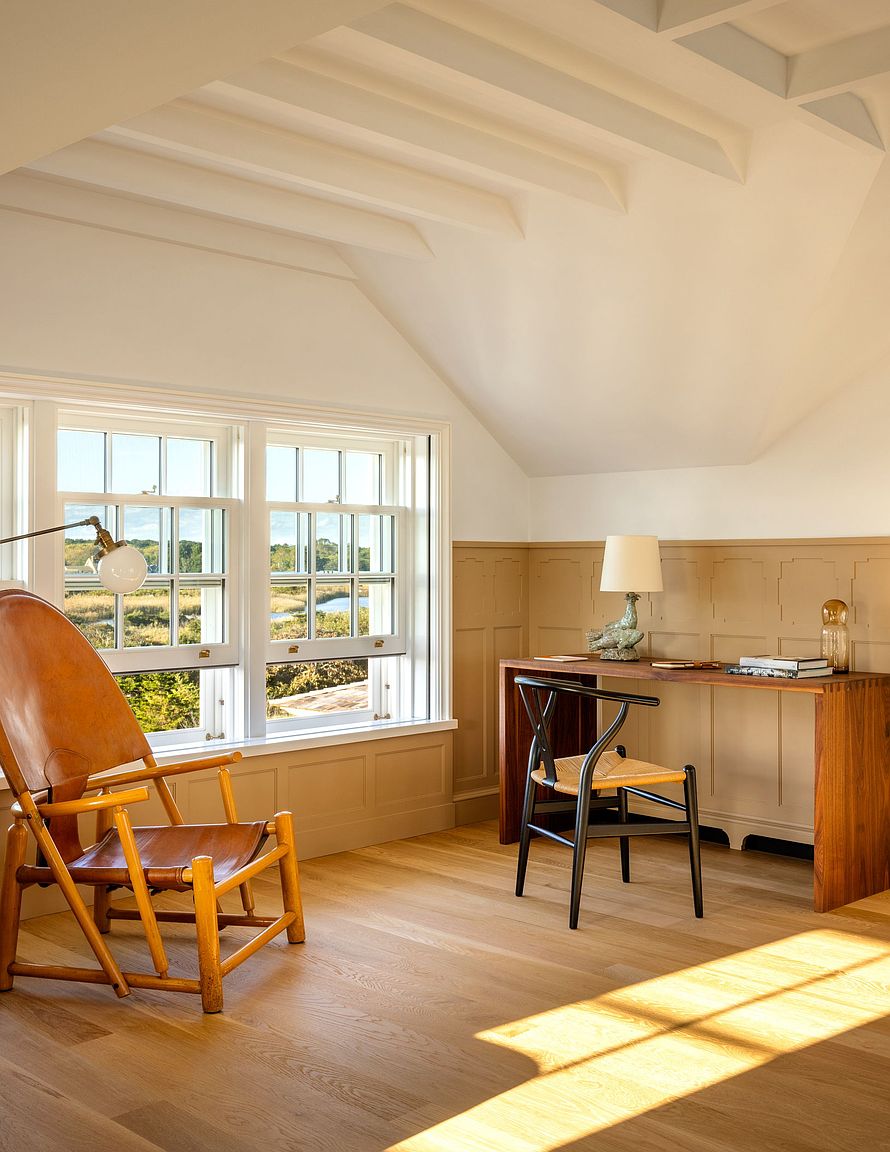 This bright and serene home office features a warm, neutral color palette with paneled wainscoting and light wood flooring. A sleek wooden desk is paired with a black wishbone-style chair, while a leather lounge chair sits near the large, multi-pane windows that offer a view of the outdoors. The vaulted ceiling with exposed beams adds architectural interest to this peaceful, sun-drenched workspace.