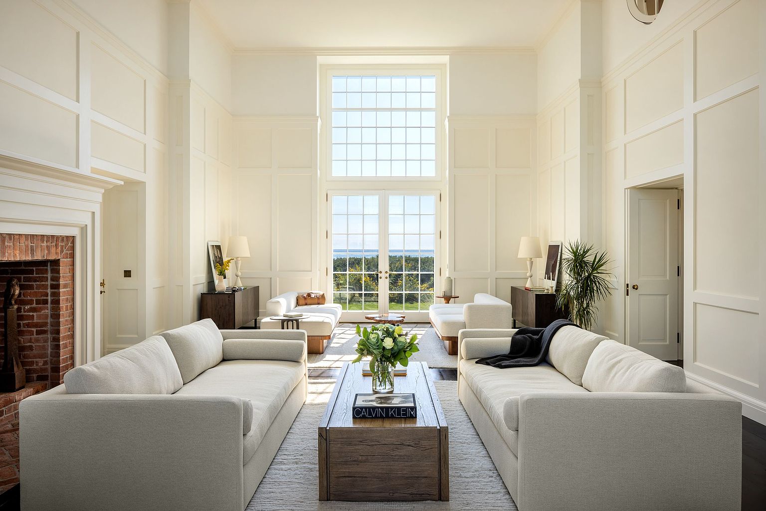 This elegant, high-ceilinged living room features a symmetrical layout with two plush, light-toned sofas facing each other across a substantial wooden coffee table. A tall, multi-paned window wall serves as the focal point, framing a serene view of the outdoors and flooding the space with natural light. To the left, a classic brick fireplace adds warmth to the room, which is finished with crisp white wall paneling and dark flooring, creating a sophisticated and airy atmosphere.