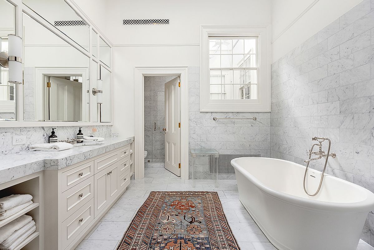 This elegant primary bathroom features a freestanding soaking tub positioned near a large window, complemented by a long vanity with stone countertops and ample storage. The space is finished with light-toned stone wall tiles and a patterned area rug that adds warmth to the bright, airy atmosphere. A doorway leads to a separate water closet, creating a well-organized and luxurious layout.