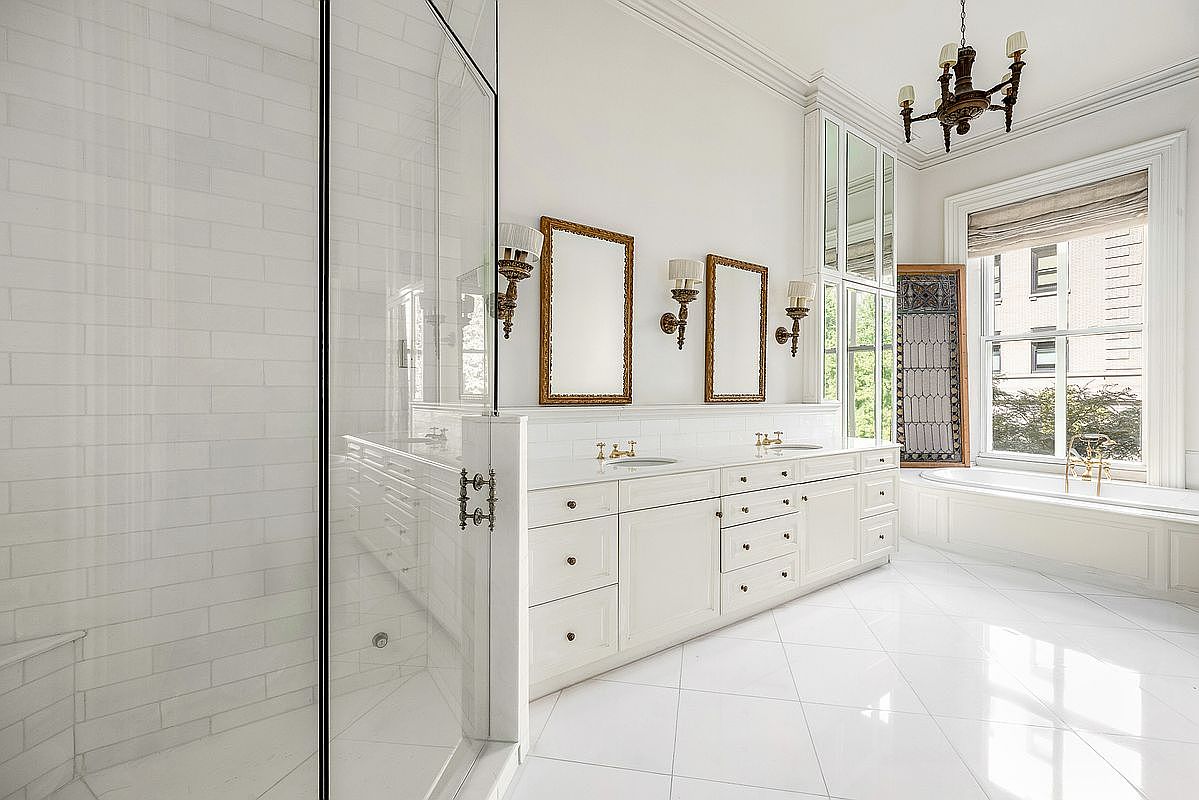 This elegant primary bathroom features a double vanity with white cabinetry, gold-toned hardware, and two rectangular mirrors framed in gold. To the left, a glass-enclosed shower with white subway tile is visible, while a deep soaking tub sits beneath a large window on the right. The room is illuminated by a decorative chandelier and wall-mounted sconces, creating a bright and sophisticated atmosphere.