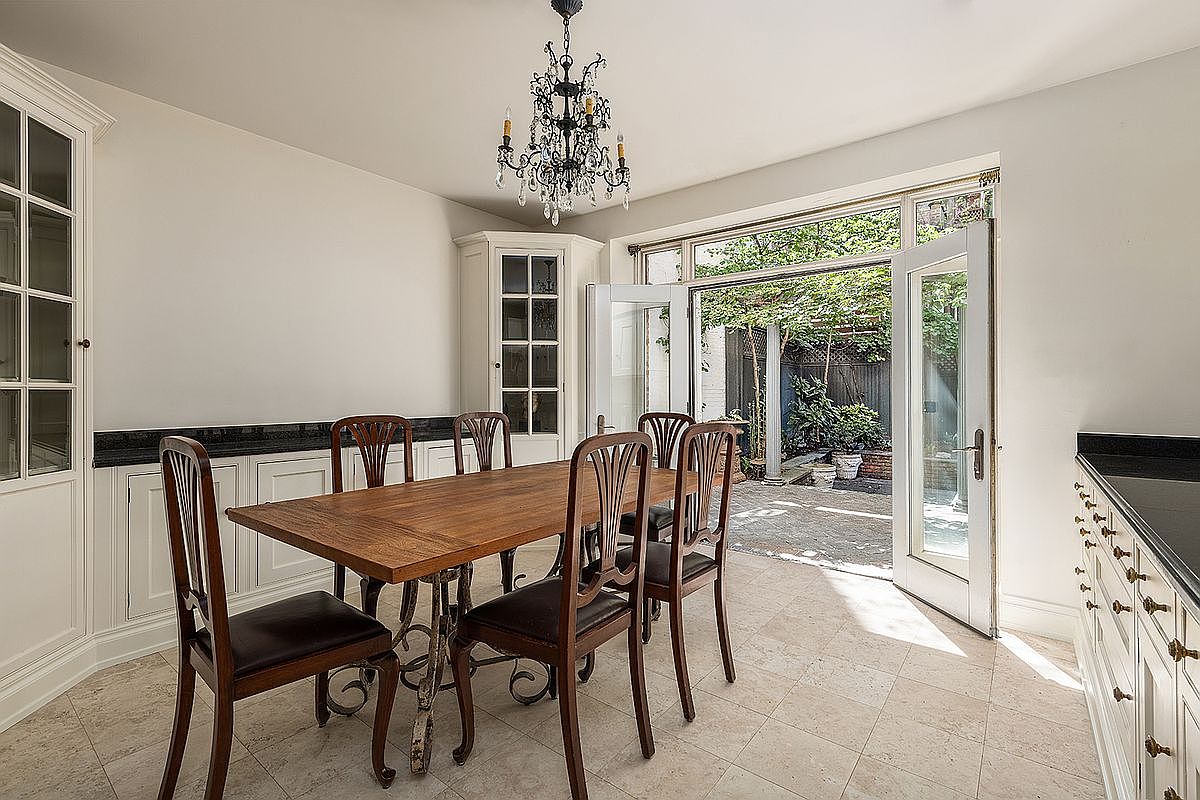 This elegant dining room features a warm wood table surrounded by four chairs, set against a backdrop of white built-in cabinetry and a classic chandelier. Large glass doors open directly onto a private, lush garden patio, creating a seamless transition between indoor and outdoor living spaces. The room is finished with light-toned tile flooring and a clean, neutral color palette that enhances the bright, airy atmosphere.