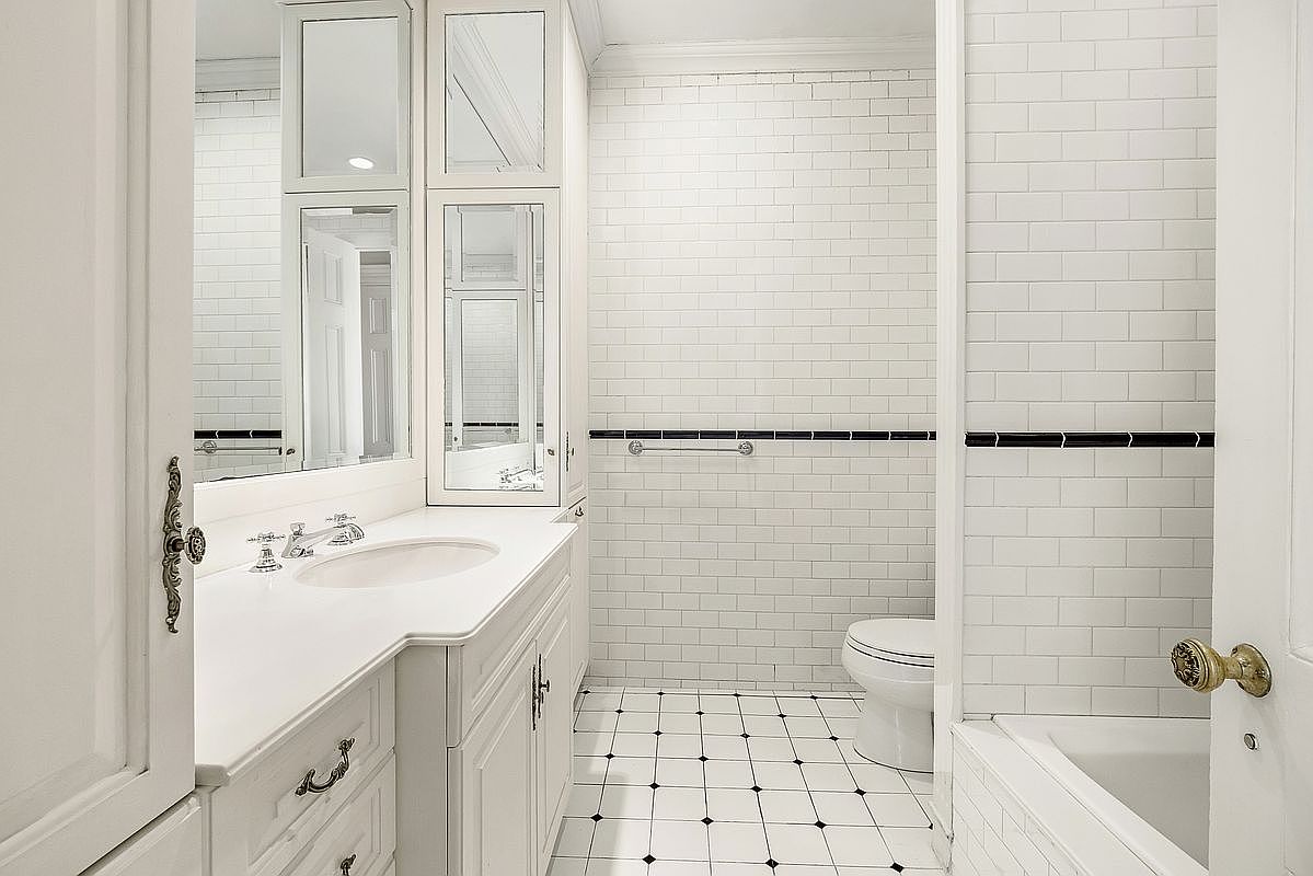 This bright, classic bathroom features white subway tile walls accented by a black horizontal border and white-and-black patterned floor tiles. The space includes a white vanity with a stone countertop, a built-in medicine cabinet with mirrored doors, and a bathtub tucked into the corner. The perspective is from the doorway, looking into the room to showcase the clean, traditional design.