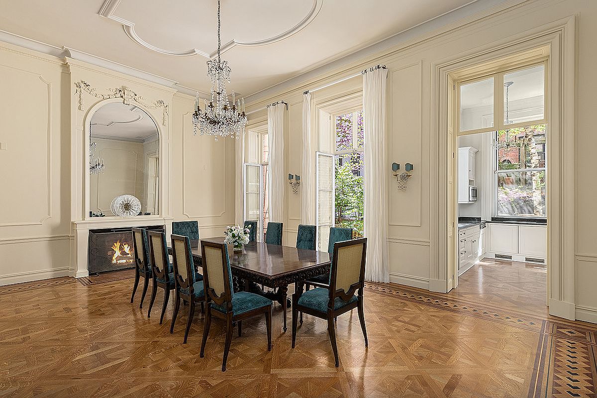 This elegant formal dining room features intricate parquet wood flooring and classic wall molding, centered around a large dark wood table with upholstered teal chairs. A crystal chandelier hangs from the ornate ceiling, while a tall fireplace surround with an integrated mirror serves as a sophisticated focal point. Large windows with sheer curtains provide natural light and a view of the greenery outside, and a wide doorway leads into an adjacent kitchen area.