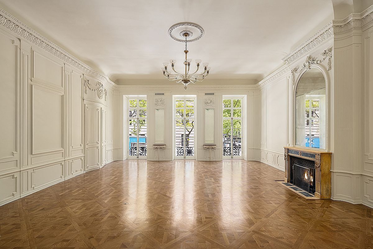 This elegant, formal living room features intricate wall paneling, ornate crown molding, and a classic herringbone-patterned wood floor. A grand crystal chandelier hangs from a decorative ceiling medallion, while three sets of tall glass-paned doors allow abundant natural light to fill the space. To the right, a fireplace with a stone mantel and a large arched mirror above serves as a sophisticated focal point in this bright, airy room.